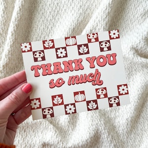 May include: A cream-colored card with the words "THANK YOU so much" in a retro font. The card features a checkered border with fall-themed illustrations of leaves, pumpkins, flowers, and mushrooms. The card is held by a person.