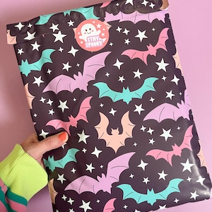 Pode incluir: Um envelope de envio com padr&atilde;o preto e branco com morcegos rosa e turquesa e estrelas brancas. O envelope tem um adesivo com um fantasma de desenho animado e o texto "Stay Spooky".