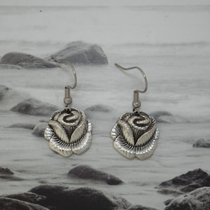 Op de afbeelding: Een paar zilverkleurige rozenoorbellen. Elke oorbel heeft een gedetailleerd rozenontwerp met een haakje voor doorboorde oren. De oorbellen worden getoond tegen een vage, grijstinten achtergrond.