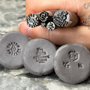 May include: Close-up of five metal stamps with different designs, including "Custom Pottery", "Ceramics Studio", and "LIP". The stamps are held in a hand, and the background is a gray, textured surface.