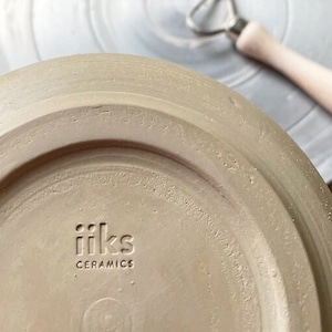 May include: Close-up of the bottom of a light brown ceramic plate with the text "iiks ceramics" stamped in black.