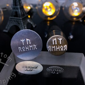 K&ouml;nnte beinhalten: Zwei Metallstempel mit gravierten Runen. Der obere Stempel ist silberfarben und tr&auml;gt die Aufschrift "Yggdrasil". Der untere Stempel ist messingfarben und tr&auml;gt die Aufschrift "Jormungandr".