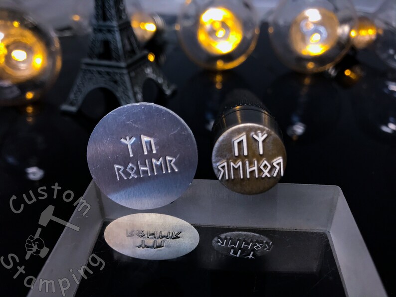 K&ouml;nnte beinhalten: Zwei Metallstempel mit gravierten Runen. Der obere Stempel ist silberfarben und tr&auml;gt die Aufschrift "Yggdrasil". Der untere Stempel ist messingfarben und tr&auml;gt die Aufschrift "Jormungandr".