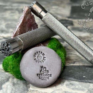 May include: A silver metal stamp with the text "CERAMIC STUDIO" and a design of three pots. The stamp is attached to a silver metal handle with a textured grip. The stamp is used to create impressions in clay. The stamp is shown next to a gray clay tile with the text "CUSTOM POTS" and "CERAMICS STUDIO" stamped into it.