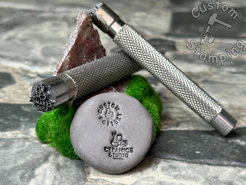 May include: A silver metal stamp with the text "CERAMIC STUDIO" and a design of three pots. The stamp is attached to a silver metal handle with a textured grip. The stamp is used to create impressions in clay. The stamp is shown next to a gray clay tile with the text "CUSTOM POTS" and "CERAMICS STUDIO" stamped into it.