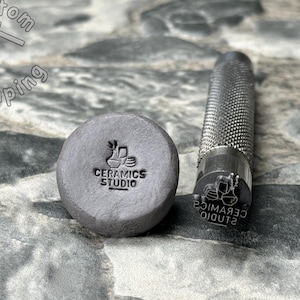 May include: A close-up of a metal stamp with the text "CERAMICS STUDIO" and a graphic of three ceramic mugs. The stamp is next to a gray clay impression of the stamp.