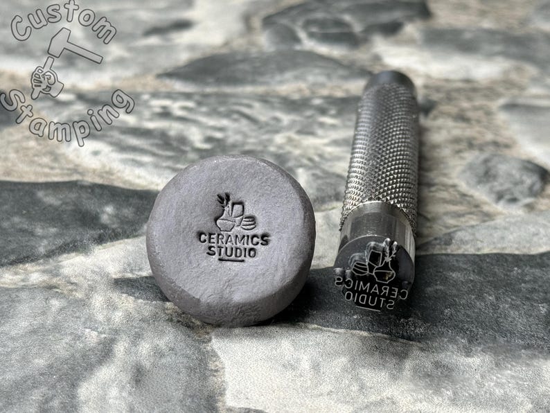 May include: A close-up of a metal stamp with the text "CERAMICS STUDIO" and a graphic of three ceramic mugs. The stamp is next to a gray clay impression of the stamp.