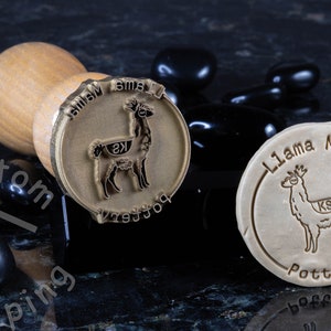 Może przedstawiać: Dwa stempla mosiężne z drewnianymi uchwytami. Stempla przedstawiają wz&oacute;r lamy z tekstem "Llama Mama Pottery" i "Llama Mama" na stemplach.