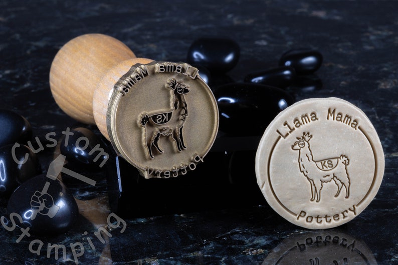 Może przedstawiać: Dwa stempla mosiężne z drewnianymi uchwytami. Stempla przedstawiają wz&oacute;r lamy z tekstem "Llama Mama Pottery" i "Llama Mama" na stemplach.