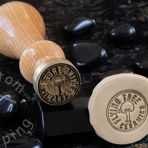 Może przedstawiać: Drewniany uchwyt przymocowany do stempla mosiężnego z napisem "Flying Tree Ceramics" i stylizowanym wzorem drzewa. Stempel znajduje się na czarnej powierzchni z czarnymi kamieniami.