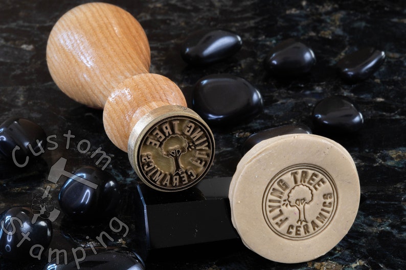 Może przedstawiać: Drewniany uchwyt przymocowany do stempla mosiężnego z napisem "Flying Tree Ceramics" i stylizowanym wzorem drzewa. Stempel znajduje się na czarnej powierzchni z czarnymi kamieniami.