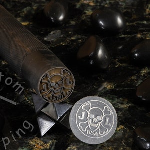 K&ouml;nnte beinhalten: Ein schwarzer Stempel aus Metall mit einem Totenkopf- und Knochenkreuz-Design, den Buchstaben "J.L." und dem Text "42U". Der Stempel befindet sich auf einer schwarzen Oberfl&auml;che mit schwarzen Steinen.