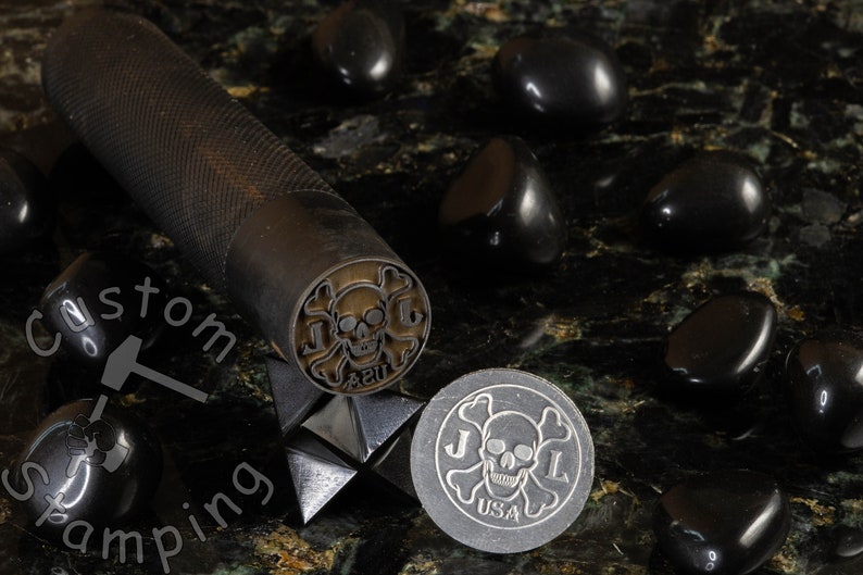 K&ouml;nnte beinhalten: Ein schwarzer Stempel aus Metall mit einem Totenkopf- und Knochenkreuz-Design, den Buchstaben "J.L." und dem Text "42U". Der Stempel befindet sich auf einer schwarzen Oberfl&auml;che mit schwarzen Steinen.