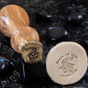 Może przedstawiać: Stempel z drewnianym uchwytem i mosiężną głowicą przedstawiający dom z dymem wydobywającym się z komina. Stempel służy do tworzenia odcisku na mydle, na kt&oacute;rym widnieje napis "House of Blue" i "Handmade".