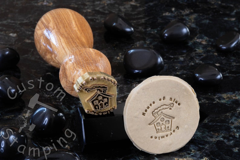 Może przedstawiać: Stempel z drewnianym uchwytem i mosiężną głowicą przedstawiający dom z dymem wydobywającym się z komina. Stempel służy do tworzenia odcisku na mydle, na kt&oacute;rym widnieje napis "House of Blue" i "Handmade".