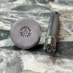 May include: A close-up of a metal stamp with the text "CUSTOM POTTERY" and a hand holding a vase. The stamp is next to a gray clay imprint of the stamp.