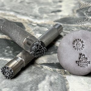 May include: Two metal stamps with handles and a gray clay tile with two impressions. The stamps are engraved with the text "Studio Ceramics" and a design of three ceramic pots. The clay tile has the text "Custom Pottery" and "Studio Ceramics" with a design of a ceramic pot.