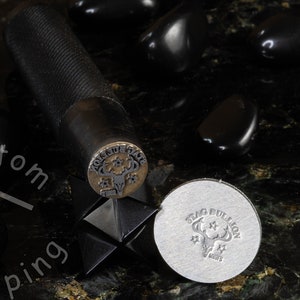 K&ouml;nnte beinhalten: Nahaufnahme eines Metallstempels mit einem Hirschkopfdesign und den Worten "Stag Bullion" darum herum. Der Stempel befindet sich auf einer schwarzen Oberfl&auml;che mit schwarzen Steinen.