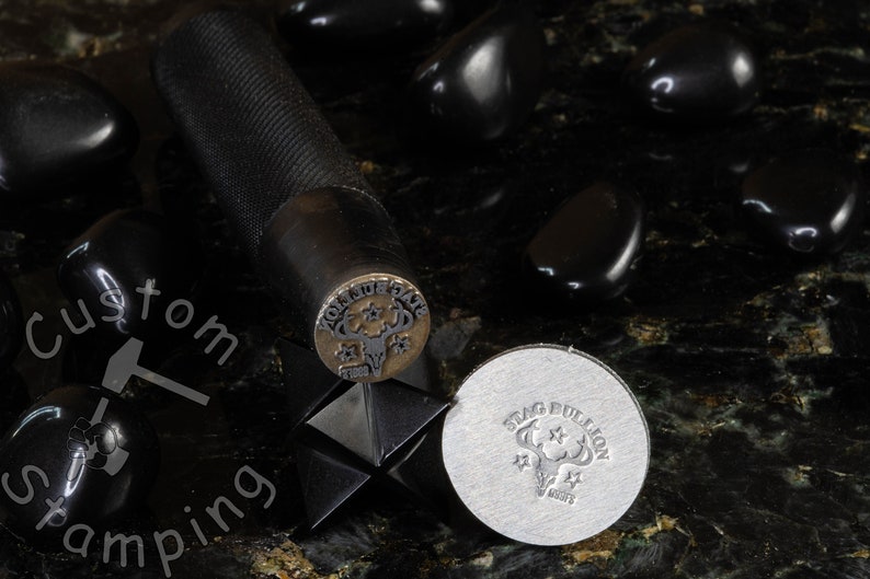 K&ouml;nnte beinhalten: Nahaufnahme eines Metallstempels mit einem Hirschkopfdesign und den Worten "Stag Bullion" darum herum. Der Stempel befindet sich auf einer schwarzen Oberfl&auml;che mit schwarzen Steinen.