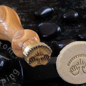 Może przedstawiać: Stempel mosiężny z drewnianym uchwytem, z tekstem "Made By Tiny Hands" i dwoma rękami z uniesionymi palcami. Stempel leży na czarnej powierzchni z czarnymi kamieniami. Stempel został użyty do stworzenia odcisku na kawałku gliny.