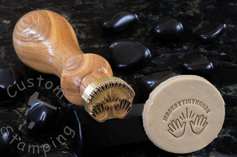 Może przedstawiać: Stempel mosiężny z drewnianym uchwytem, z tekstem "Made By Tiny Hands" i dwoma rękami z uniesionymi palcami. Stempel leży na czarnej powierzchni z czarnymi kamieniami. Stempel został użyty do stworzenia odcisku na kawałku gliny.
