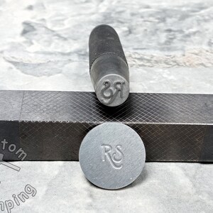 K&ouml;nnte beinhalten: Nahaufnahme eines Metallstempels mit den eingravierten Buchstaben "RS". Der Stempel liegt auf einem schwarzen Metallblock mit einem Rautenmuster. Der Stempel wird verwendet, um individuelle Designs auf Metall zu erstellen.