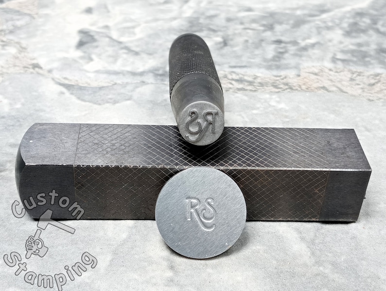 K&ouml;nnte beinhalten: Nahaufnahme eines Metallstempels mit den eingravierten Buchstaben "RS". Der Stempel liegt auf einem schwarzen Metallblock mit einem Rautenmuster. Der Stempel wird verwendet, um individuelle Designs auf Metall zu erstellen.