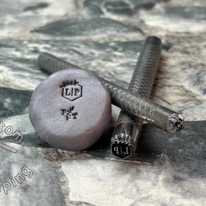 May include: Close-up of three metal stamps with the letters "LIP" and a small leaf design. The stamps are on a gray textured surface.