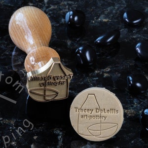 Może przedstawiać: Stempel mosiężny z tekstem "Silenced Vessel Waffle-the" i stylizowanym obrazem wazonu i miski. Stempel znajduje się obok brązowego glinianego odcisku stempla z tekstem "Tracey DeLellis art-pottery".