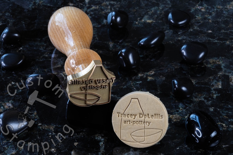 Może przedstawiać: Stempel mosiężny z tekstem "Silenced Vessel Waffle-the" i stylizowanym obrazem wazonu i miski. Stempel znajduje się obok brązowego glinianego odcisku stempla z tekstem "Tracey DeLellis art-pottery".