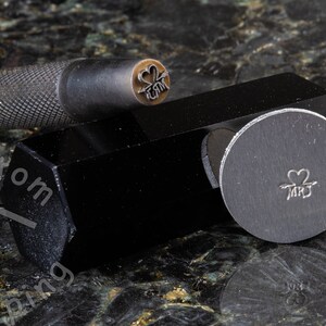 K&ouml;nnte beinhalten: Nahaufnahme eines Metallstempels mit einem Herz- und Pfeilmotiv und dem Text "MR & MRS". Der Stempel befindet sich auf einer schwarzen Metallbasis.