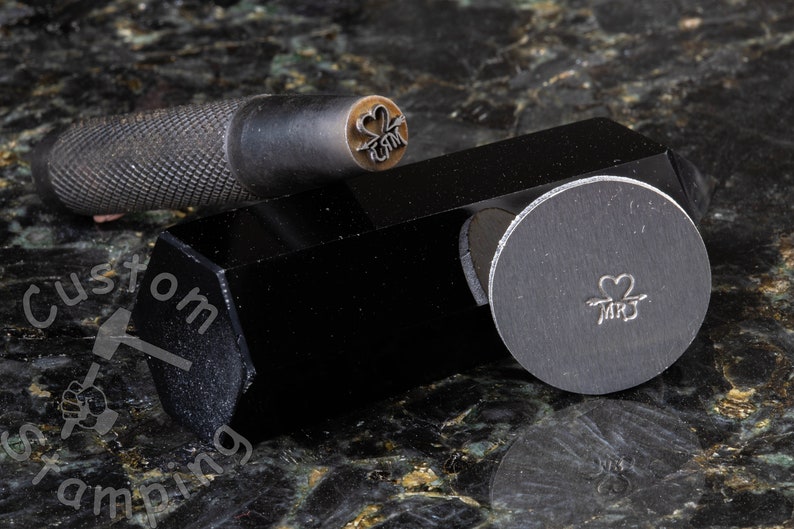 K&ouml;nnte beinhalten: Nahaufnahme eines Metallstempels mit einem Herz- und Pfeilmotiv und dem Text "MR & MRS". Der Stempel befindet sich auf einer schwarzen Metallbasis.