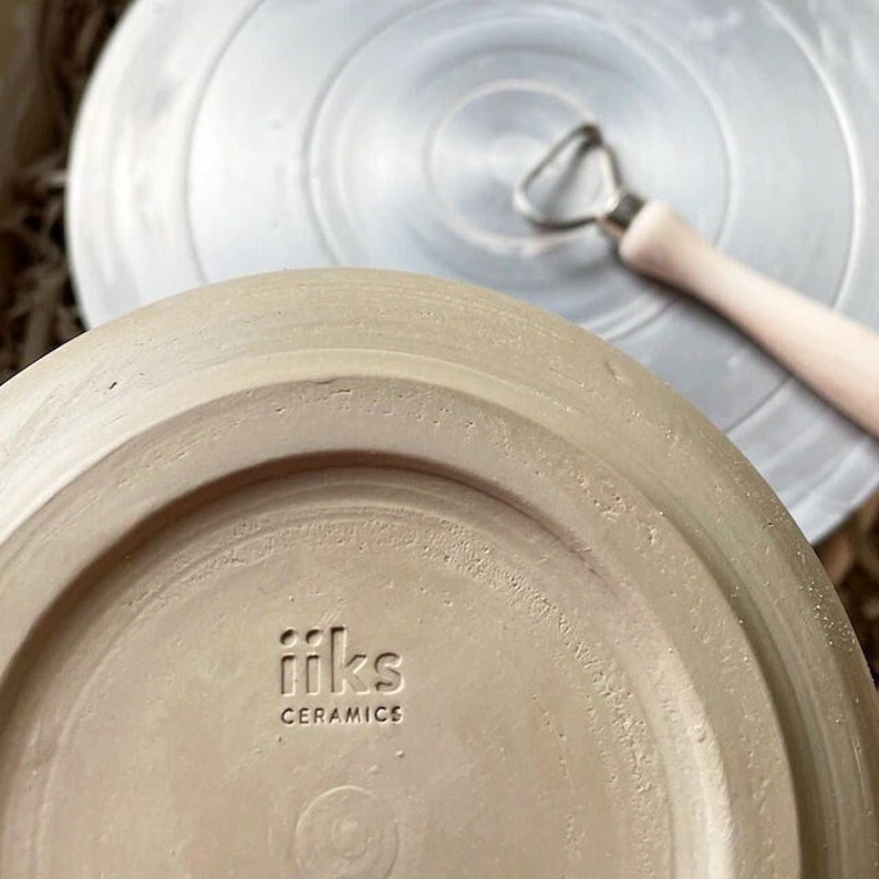 Może przedstawiać: Zbliżenie na sp&oacute;d jasnobrązowego talerza ceramicznego z wytłoczonym czarnym tekstem "iiks ceramics".