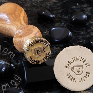 Może przedstawiać: Drewniany uchwyt przymocowany do stempla mosiężnego z tekstem "HANDCRAFTED BY SHARI ORBACH" i stylizowanym obrazem filiżanki do kawy. Stempel znajduje się na brązowej powierzchni z czarnymi kamieniami.