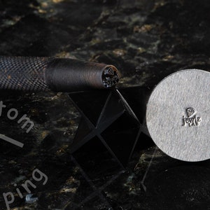 K&ouml;nnte beinhalten: Ein Metallstempelwerkzeug f&uuml;r Schmuck mit einem Diamantendesign. Das Werkzeug befindet sich auf einer schwarzen Oberfl&auml;che mit einem schwarz-wei&szlig;en Logo, auf dem "Custom Stamping" steht.