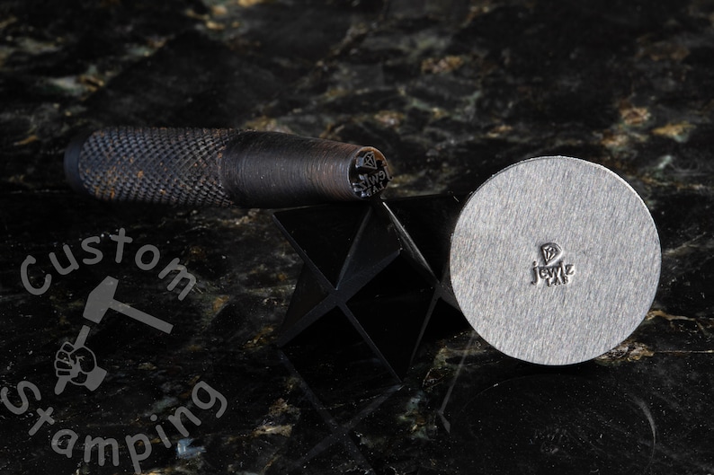 K&ouml;nnte beinhalten: Ein Metallstempelwerkzeug f&uuml;r Schmuck mit einem Diamantendesign. Das Werkzeug befindet sich auf einer schwarzen Oberfl&auml;che mit einem schwarz-wei&szlig;en Logo, auf dem "Custom Stamping" steht.