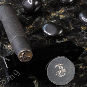 K&ouml;nnte beinhalten: Ein schwarzer Stempel aus Metall mit einem detaillierten Design einer Schildkr&ouml;te. Der Stempel befindet sich auf einer schwarzen Oberfl&auml;che mit schwarzen Steinen drumherum. Der Stempel ist mit "JOHAVI TURTLE" beschriftet.