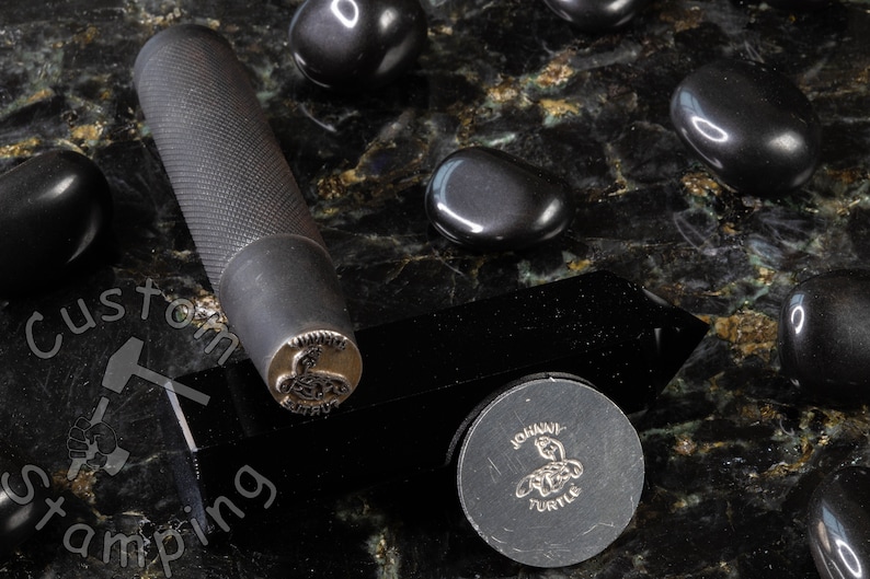 K&ouml;nnte beinhalten: Ein schwarzer Stempel aus Metall mit einem detaillierten Design einer Schildkr&ouml;te. Der Stempel befindet sich auf einer schwarzen Oberfl&auml;che mit schwarzen Steinen drumherum. Der Stempel ist mit "JOHAVI TURTLE" beschriftet.