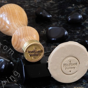 Może przedstawiać: Drewniany stempel z mosiężną głowicą, z tekstem "Re-Root Pottery" w okrągłym wzorze. Stempel leży na czarnej powierzchni obok odcisku gliny w kolorze beżowym.