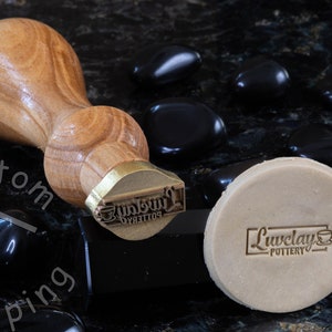 Może przedstawiać: Stempel z drewnianym uchwytem i mosiężną głowicą. Stempel jest grawerowany tekstem "Liveclay Pottery". Stempel leży na kawałku gliny z tym samym tekstem odciśniętym na nim.