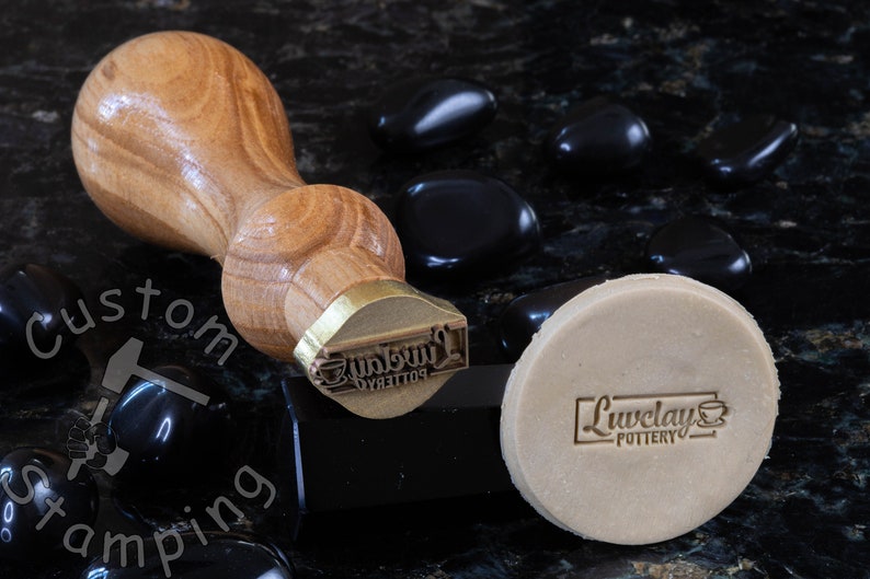 Może przedstawiać: Stempel z drewnianym uchwytem i mosiężną głowicą. Stempel jest grawerowany tekstem "Liveclay Pottery". Stempel leży na kawałku gliny z tym samym tekstem odciśniętym na nim.