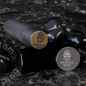 K&ouml;nnte beinhalten: Nahaufnahme eines Metallstempels mit dem Text "HARVEE ROSE" und einem Rosenmotiv. Der Stempel befindet sich auf einer schwarzen Oberfl&auml;che mit schwarzen Steinen.