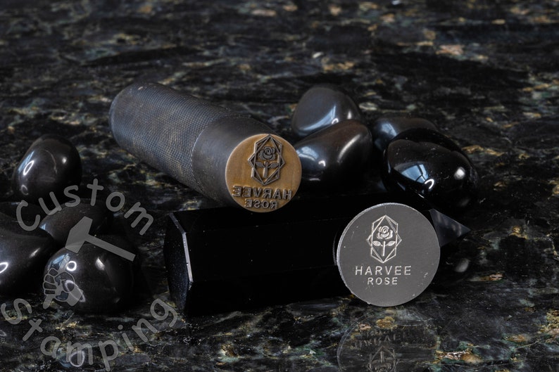 K&ouml;nnte beinhalten: Nahaufnahme eines Metallstempels mit dem Text "HARVEE ROSE" und einem Rosenmotiv. Der Stempel befindet sich auf einer schwarzen Oberfl&auml;che mit schwarzen Steinen.