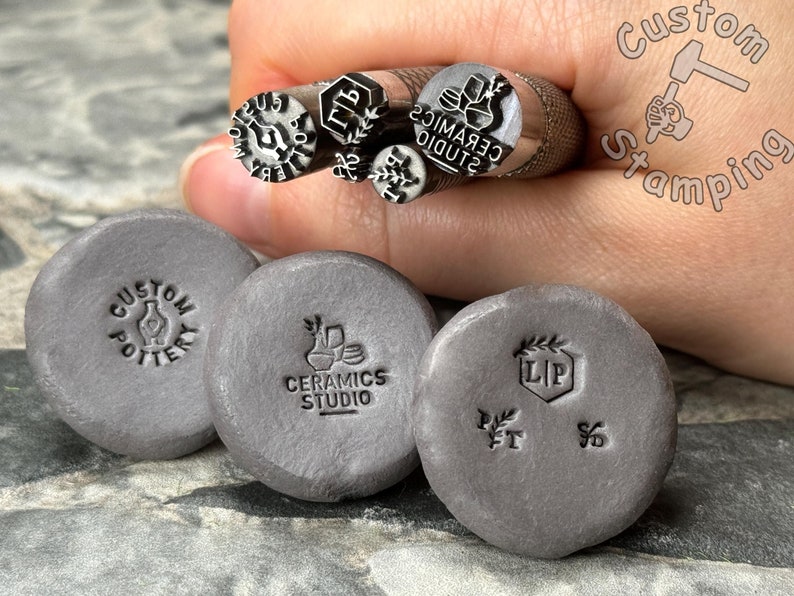 May include: Close-up of five metal stamps with different designs, including "Custom Pottery", "Ceramics Studio", and "LIP". The stamps are held in a hand, and the background is a gray, textured surface.