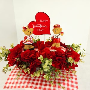 May include: A red and white checkered tablecloth with a red heart that says "Happy Valentine's Day" and two teddy bears. One teddy bear is wearing a red and white dress and the other is wearing a red and white uniform. The teddy bears are surrounded by red flowers.