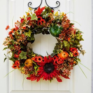 May include: A fall-themed wreath with red, orange, and yellow flowers, green leaves, and small pumpkins. The wreath is hanging on a white door.