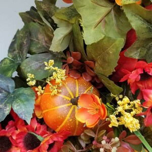 May include: A fall-themed wreath with orange and red flowers, green leaves, and a small orange pumpkin.