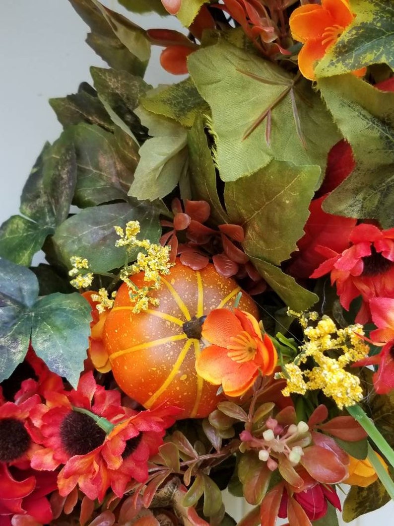 May include: A fall-themed wreath with orange and red flowers, green leaves, and a small orange pumpkin.