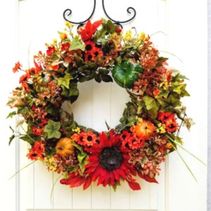 May include: A fall-themed wreath with red, orange, and yellow flowers, green leaves, and small gourds. The wreath is decorated with a large sunflower in the center.