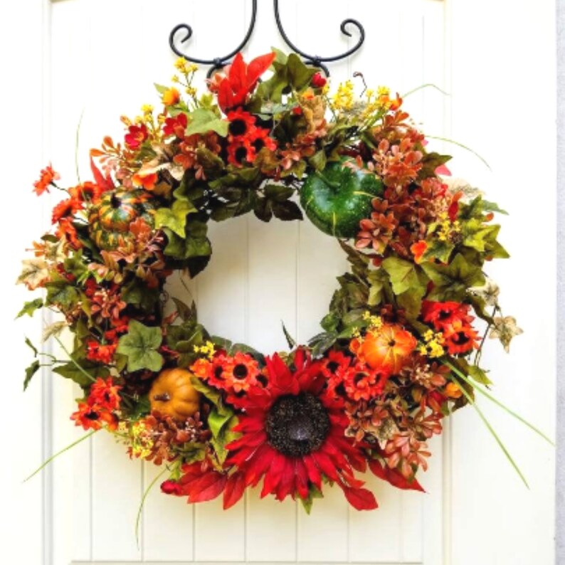 May include: A fall-themed wreath with red, orange, and yellow flowers, green leaves, and small gourds. The wreath is decorated with a large sunflower in the center.